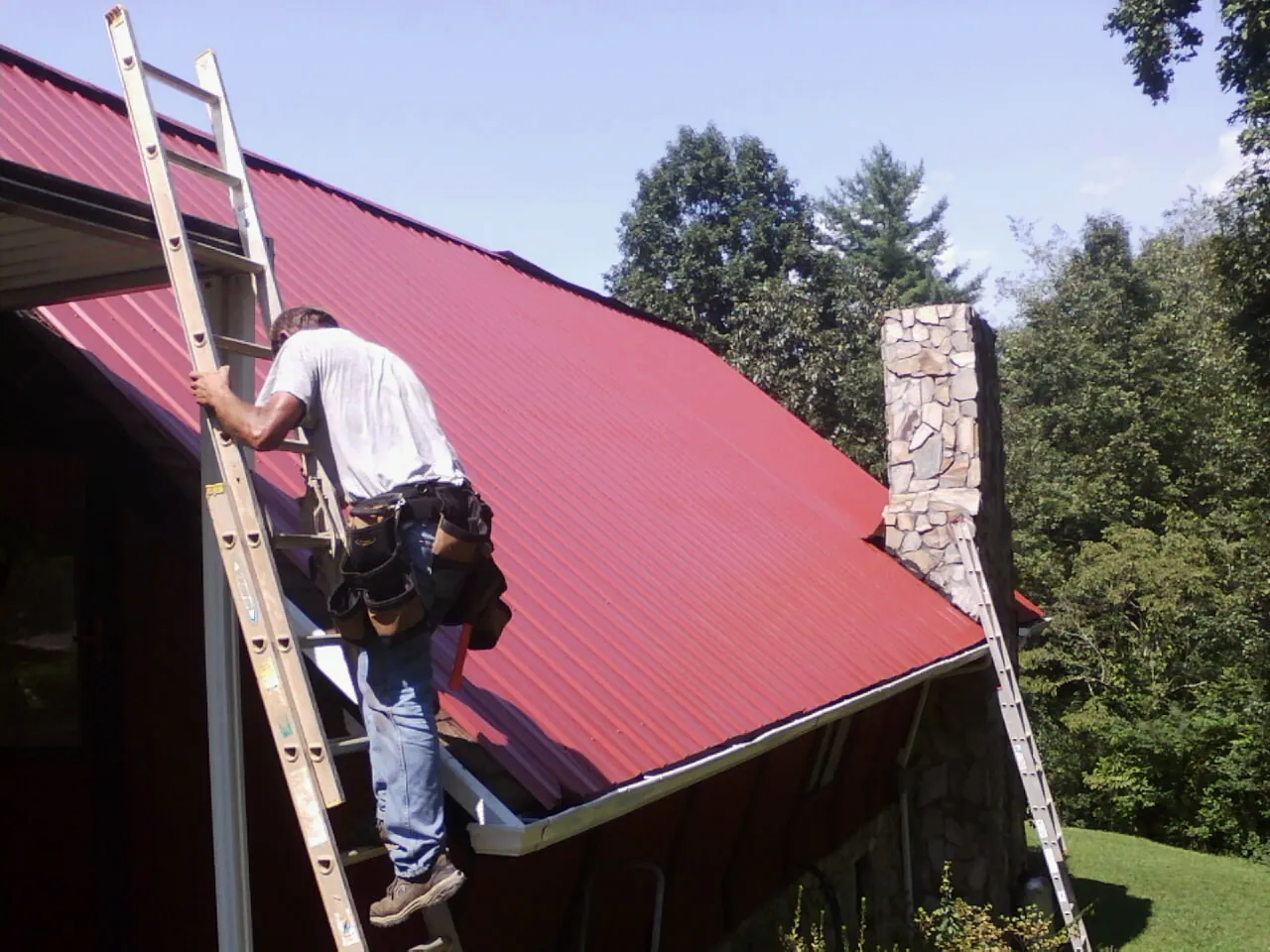 Roofer on a ladder beside a red metal roof and stone chimney — Soffit & Fascia Repair by Roofing Brothers in Wolf Creek